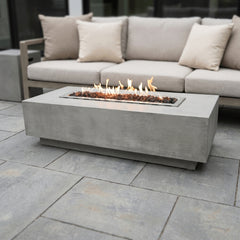 Light gray Elementi Granville fire pit table with flames lit beside an outdoor sofa on a daylight patio