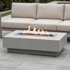Light gray Elementi Hampton fire pit table with flames lit beside an outdoor sofa on a daylight patio