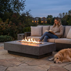 Woman reading beside the low 14-inch-tall Elementi Hampton fire pit table at dusk with a dog resting nearby