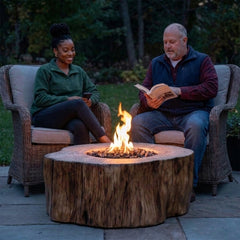 Two friends relaxing in wicker chairs beside the 17-inch-tall Elementi Manchester stump fire table at dusk