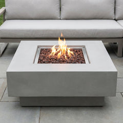Light gray Elementi Manhattan square fire pit table with flames lit on a stone patio in daylight