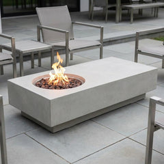 Light gray Elementi Metropolis fire pit table with flames lit on a stone patio in daylight