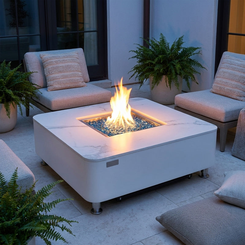 Elementi Plus Annecy square white porcelain fire table with round flame over Caribbean blue fire glass in a courtyard at dusk
