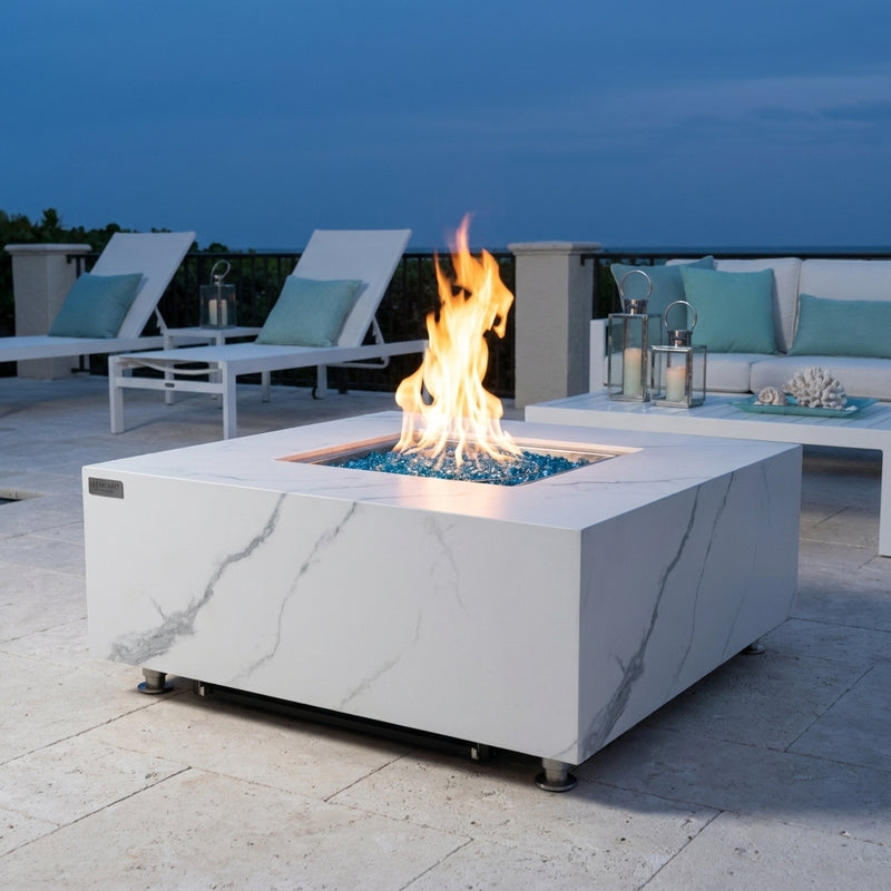 Elementi Plus Bianco white marble porcelain fire table with flames over Caribbean blue fire glass at dusk