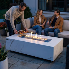 Host serving a charcuterie board on the 17-inch-tall Elementi Plus Carrara porcelain fire pit table at dusk