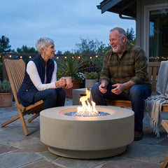 Couple seated around the knee-height Elementi Plus Colosseo round fire pit table at blue hour