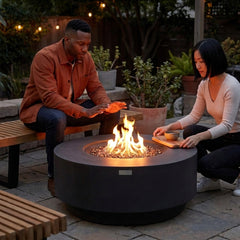Two people relaxing around the knee-height Elementi Plus Nimes round fire pit table at dusk
