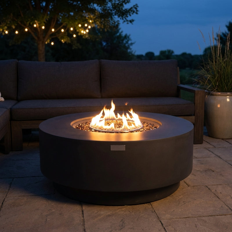Elementi Plus Nimes round dark gray concrete fire pit table with flame over bronze fire glass on a stone patio at dusk