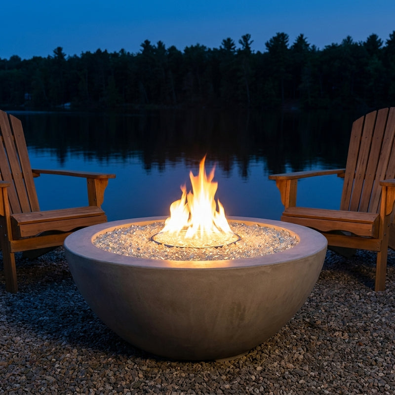 Outdoor Greatroom Cove 42 natural gray round gas fire bowl burning on a lakeside patio at dusk